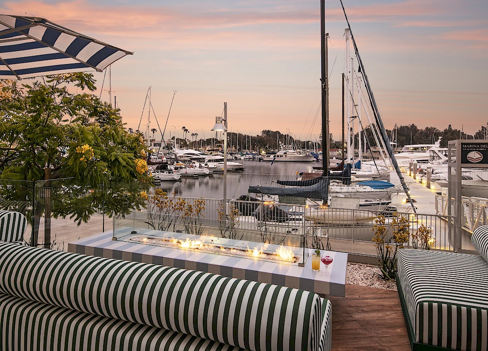 A stylish marina with boats docked, a striped outdoor seating area, a warm sunset, and soft lighting along the pier, creating a cozy waterfront vibe.